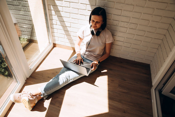 A woman is sitting on the floor near the window wearing headphones around the neck and a laptop on her feet. Top view - Foto, afbeelding
