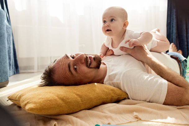 Happy dad holds his newborn baby in his arms while lying on the floor in a bright childs room. The father looks at the child with a loving look. Happy childhood and fatherhood - Zdjęcie, obraz