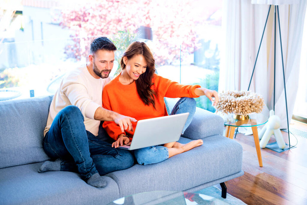 Happy couple relaxing on the couch together. Cheerful woman and handsome man using laptop while browsing on the internet. Man showing something on the screen with his finger.  - Zdjęcie, obraz