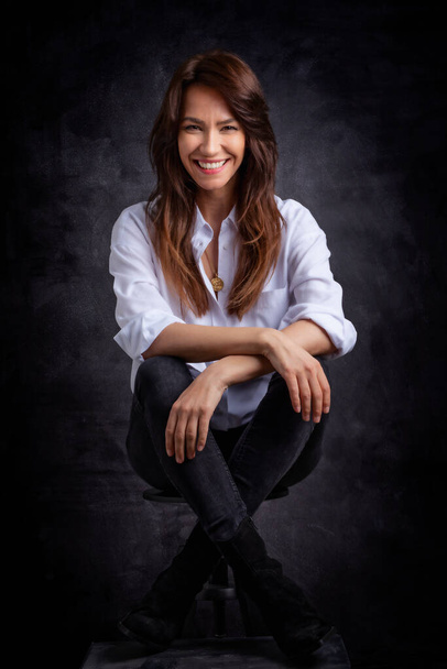 Attraktive Frau mittleren Alters mit zahmem Lächeln trägt weißes Hemd und schwarze Jeans, während sie vor einem isolierten dunklen Hintergrund sitzt. Kopierraum. Studioaufnahme. - Foto, Bild