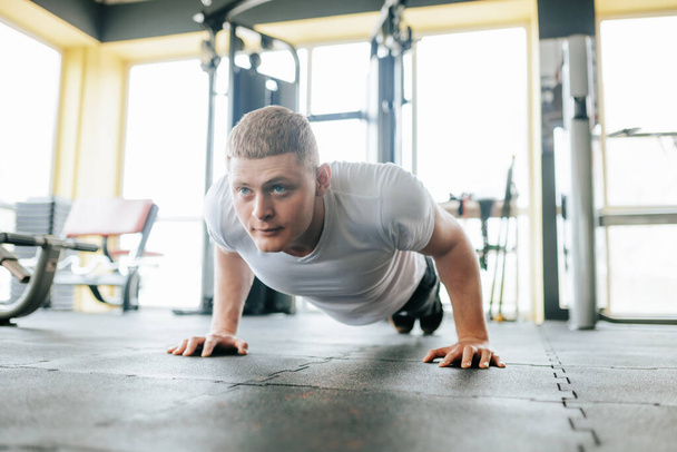 Egy határozott fiatalember fekvőtámaszozik egy jól felszerelt tornateremben az intenzív edzése alatt. Erős és sportos fizikuma megmutatja az erejét és erejét, amit kifejlesztett. - Fotó, kép