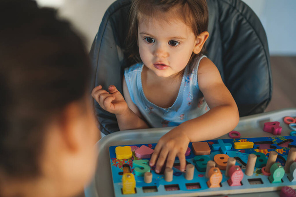 Милая маленькая девочка сидит на стульчике в гостиной дома, играя с деревянными цифрами и буквами кубики вместе со своей матерью, развитие - Фото, изображение