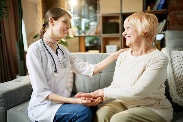 Smiling young woman, doctor supporting her elderly female patient, holding her hand and cheering up. Senior lady at home. Concept of medical care, medicine, illness, health care, profession - Фото, изображение