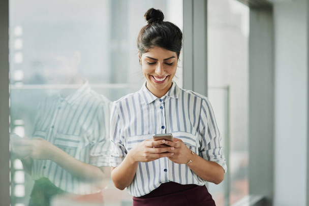 Zakelijke vrouw, glimlach en telefoon tekst met geluk op kantoor venster met een e-mail. Jonge vrouwelijke werknemer en mobiele verbinding van een werknemer die zich gelukkig voelt op sociale media en technologie in het bedrijf. - Foto, afbeelding