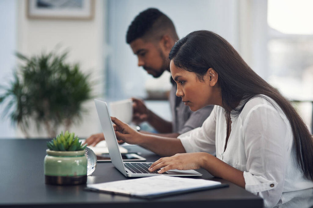 Она уделяет пристальное внимание каждой детали. молодая деловая женщина, работающая на ноутбуке в офисе со своим коллегой на заднем плане - Фото, изображение