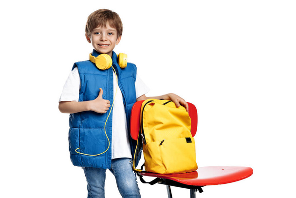 Gleeful little boy with headphone and backpack stay near red chair on white background. Back to School. Show thumb up - Photo, image