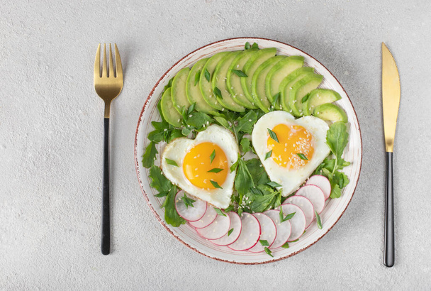 Υγιεινό ρομαντικό πρωινό - τηγανητά αυγά σε σχήμα καρδιάς σερβιρισμένα με αβοκάντο και ραπανάκι, Top view - Φωτογραφία, εικόνα