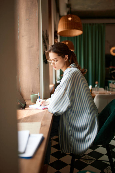 Beautiful freelancer, woman wearing eyeglasses using tablet sitting at cafe table. Young girl working online or studying. Freelance work, business people, remote job, career growth, ad concept - 写真・画像