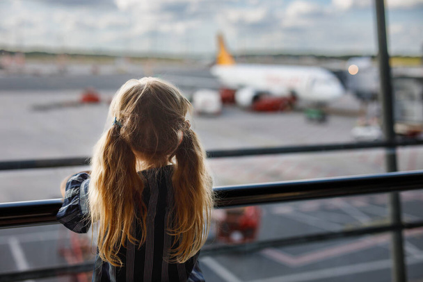 Little girl at the airport is waiting for landing at the big window. Cute kid stands at the window against the backdrop of airplanes. Looking forward to leaving for a family summer vacation - Фото, зображення