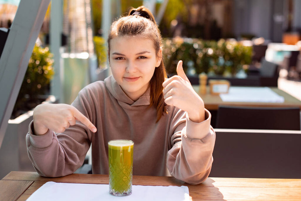 Teini-ikäinen tyttö juo lasillinen vihreää tuoretta kahvilassa kesällä aamulla ja peukalo pystyyn. keskittyä tyttö kasvot - Valokuva, kuva