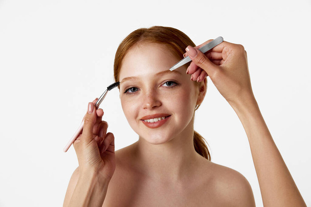 At cosmetologist. Female hands using brush and tweezer, taking care after brows of pretty young girl against white studio background. Natural beauty, makeup, cosmetology, cosmetics, skin care concept - Photo, Image