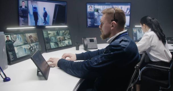 Male security officer touches tablet and controls CCTV cameras with AI face scanning. Computer monitors and big digital screens showing security cameras footage. Female colleague works at background. - Photo, Image