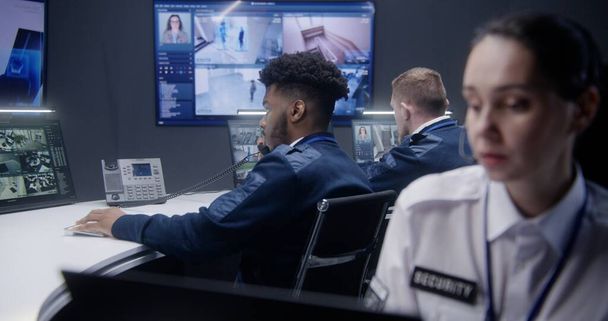 African American male security operator talks with colleague by phone in police monitoring center. Woman looks at monitors with displayed CCTV cameras view. Big screens on wall shows cameras footage. - Photo, Image