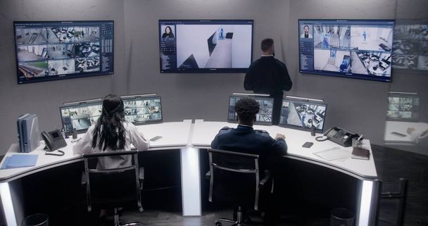 Oficiales de seguridad multiétnicos monitorean cámaras de CCTV con escaneo facial AI. Múltiples pantallas grandes con cámaras de seguridad mostradas. Trabajo en equipo en la sala de vigilancia. Seguimiento, seguimiento y seguridad social. - Foto, imagen