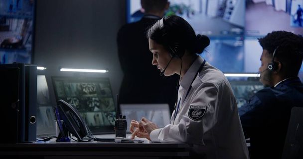 Female security officer works on computer in police monitoring center. Multi ethnic workers control surveillance CCTV cameras with tracking AI face recognition system on tablet and big digital screen. - Photo, Image