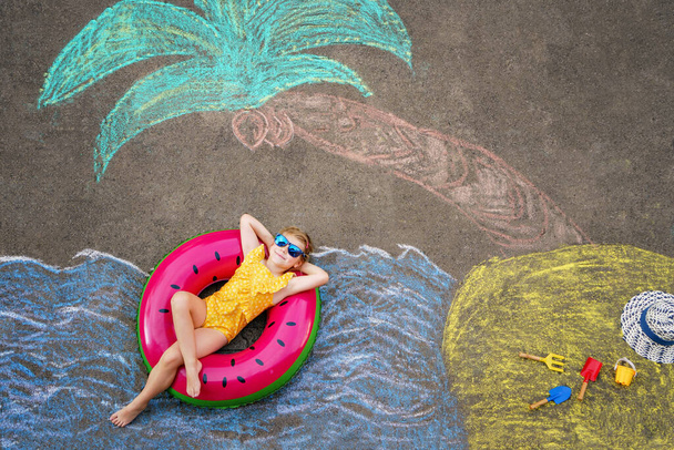 Happy little preschool girl in swimsuit on inflatable ring with sea, sand, palm painted with colorful chalks on asphalt. Cute child with having fun with chalk picture. Summer, vacations, summertime. - Foto, imagen