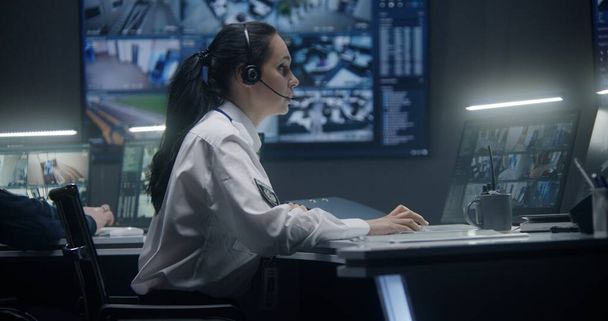 Женщина оператор в гарнитуре типов клавиатуры и контролирует камеры безопасности с помощью ИИ сканирования лица. Компьютерные мониторы и большие цифровые экраны на стене показывают записи камер наблюдения. Работа в центре наблюдения. - Фото, изображение