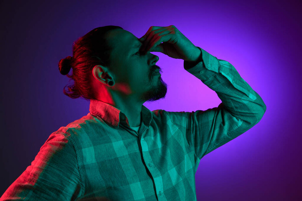 Portrait of man in checkered shirt holding head with thoughtful expression against dark gradient studio background in neon light. Concept of human emotions, facial expression, lifestyle - Foto, Bild