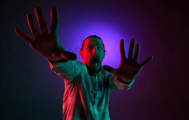 Portrait of emotional young man showing fearful face against dark gradient studio background in neon light. Horror. Concept of human emotions, facial expression, lifestyle - Foto, imagen