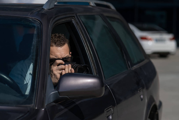 Pivate Detektiv mit Sonnenbrille, sitzt in einem Auto und führt Überwachung durch, macht Fotos mit einer professionellen Kamera - Foto, Bild