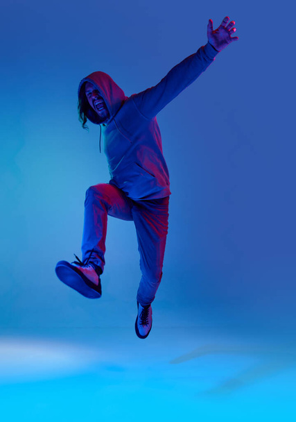 Full-length portrait of emotional man in casual clothes with hoodie jumping against blue studio background in neon light. Concept of human emotions, facial expression, lifestyle, success - Foto, Imagen