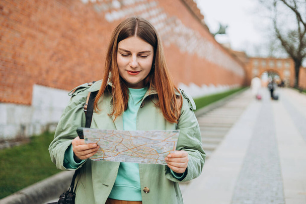 Glückliche rothaarige Frau lächelt zuversichtlich und hält Stadtplan an Straße. Attraktive junge Touristinnen erkunden die neue Stadt. Europa im Frühling bereisen. Banner des urbanen Lebensstils - Foto, Bild
