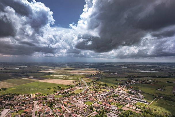 Aerial view of threatening storm clouds over a quaint village in the lush countryside of the Po Valley, Northern Italy - Фото, изображение
