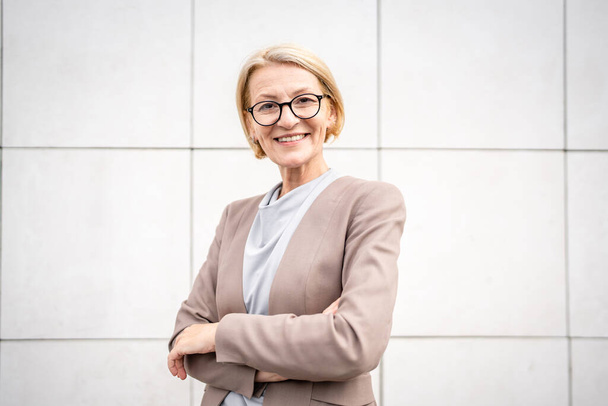 portrait of one caucasian mature woman female businesswoman blonde hair and eyeglasses stand confident outdoor in front of white wall of modern building in day copy space - Zdjęcie, obraz
