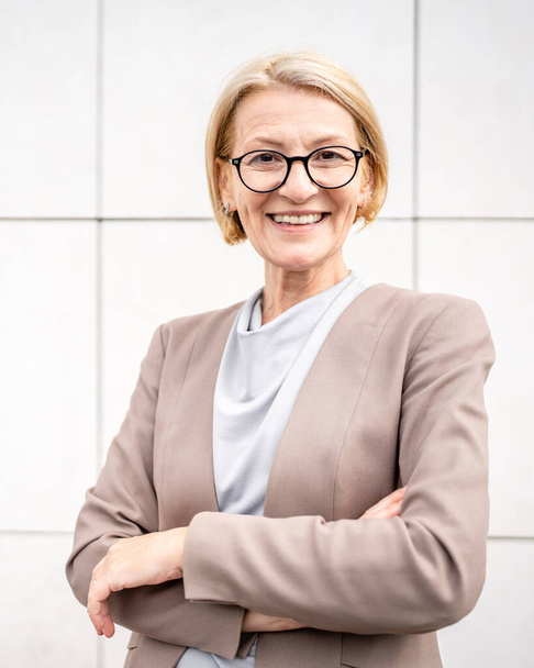 portrait of one caucasian mature woman female businesswoman blonde hair and eyeglasses stand confident outdoor in front of white wall of modern building in day copy space - Foto, Imagen