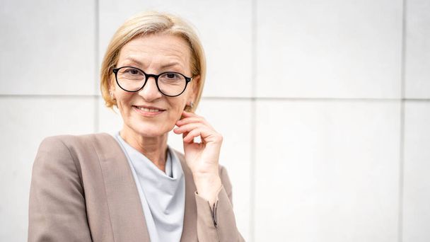 portrait of one caucasian mature woman female businesswoman blonde hair and eyeglasses stand confident outdoor in front of white wall of modern building in day copy space - Foto, immagini