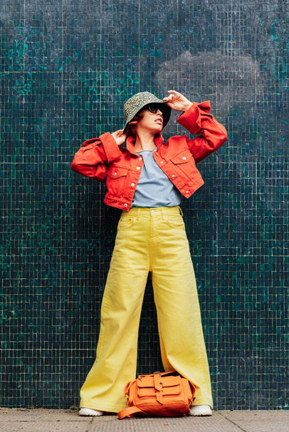 Hipster young woman in bright clothes, sun glasses, backpack bag and bucket hat posing on green blue tile wall background. Urban city street fashion. Fashion blogger. Vertical card. Selective focus - Фото, зображення