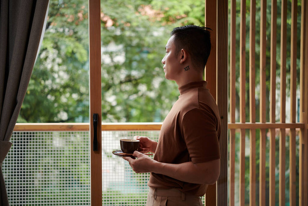 Pensive young man standing on balcony, drinking morning coffee and looking at street - Fotoğraf, Görsel