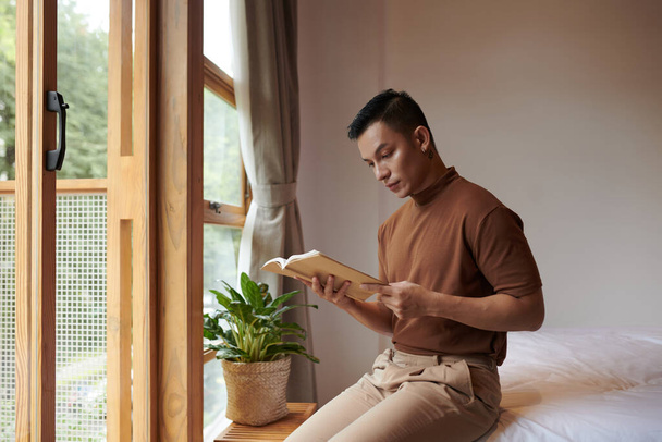 Серьезный молодой азиат, сидящий на кровати и читающий книгу рассказов - Фото, изображение