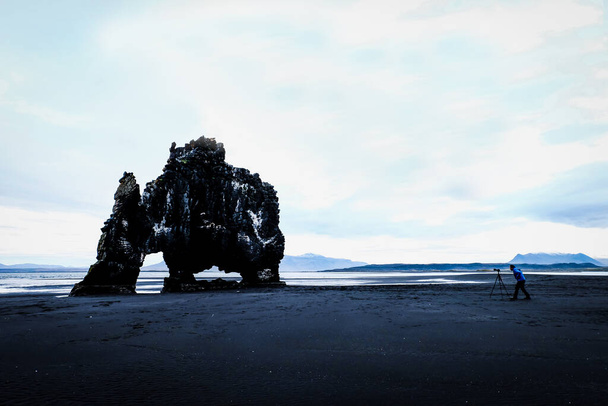Landscape of hvitserkur, acient elephant shape or dinosor aganst black sand and gray sky with little blue clound with photograper taking photo with stand. Iceland landmark destiny. - Фото, зображення