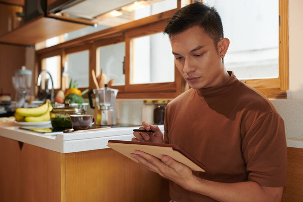 Серьезный молодой человек стоит на кухне и проверяет список продуктов на планшетном компьютере - Фото, изображение