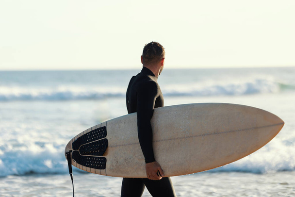 Surfer w mokrym kostiumie spacerujący po brzegu trzymający deskę surfingową i patrzący na morze. Środek strzału - Zdjęcie, obraz