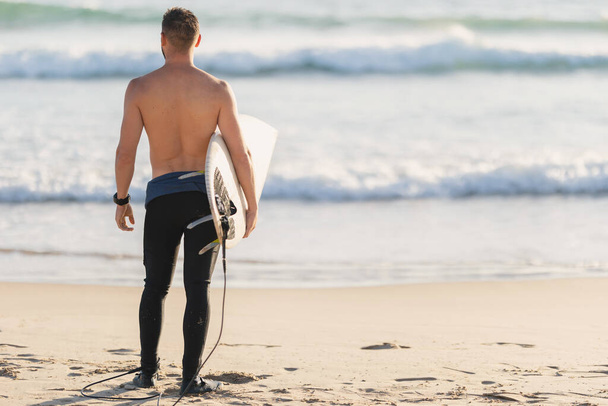 Ένας αθλητικός σέρφερ με γυμνό κορμό που στέκεται δίπλα στον ωκεανό - πίσω όψη. Μεσαίο πλάνο - Φωτογραφία, εικόνα