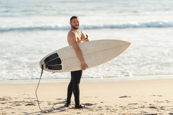 Sportowy surfer z nagim tułowiem stojący nad morzem i pokazujący kciuk w górze patrzący w kamerę. Środek strzału - Zdjęcie, obraz