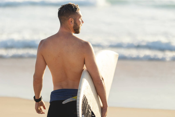 An athletic man surfer with naked torso standing by the ocean. Mid shot - Zdjęcie, obraz