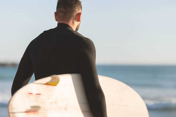 An athletic man surfer in a wetsuit standing by the ocean. Mid shot - Foto, Imagem