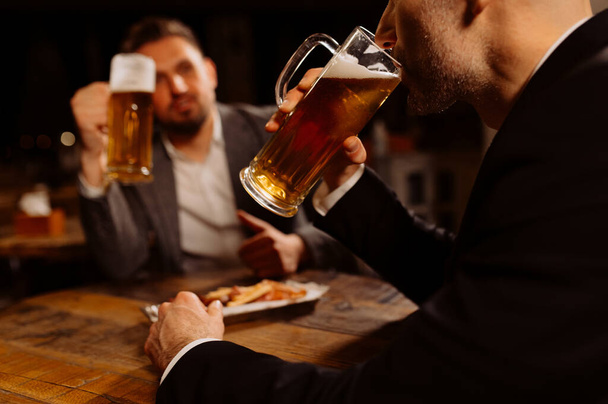Aikuiset ystävät istuvat olutmukien kanssa pubissa. Kaksi iloinen kaverit juovat luonnos olutta, juhlii kokousta ja hymyilee. - Valokuva, kuva