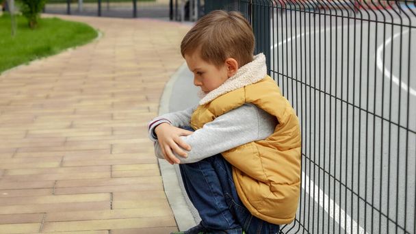 Unhappy lonely boy leaning on metal fence at school playground and sitting down. Child depression, problems with bullying, victim in school, emigration, criminal and poverty. - Photo, Image
