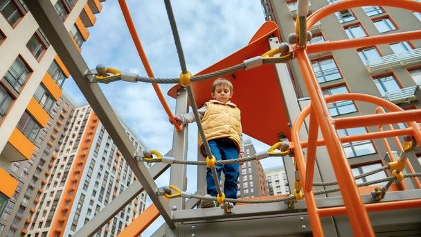 Widok z ziemi na chłopca idącego przez most linowy na placu zabaw. Aktywne dziecko, sport i rozwój, dzieci bawiące się na świeżym powietrzu. - Zdjęcie, obraz