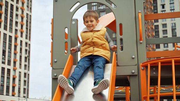 Powolny ruch wesołego chłopca zjeżdżającego ze zjeżdżalni na nowym placu zabaw. Aktywne dziecko, sport i rozwój, dzieci bawiące się na świeżym powietrzu. - Zdjęcie, obraz