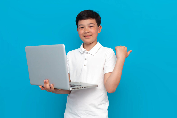 маленький азиатский мальчик, использующий ноутбук и показывающий на копировальном пространстве на синем фоне, корейский ребенок, печатающий онлайн на компьютере на изолированном фоне - Фото, изображение