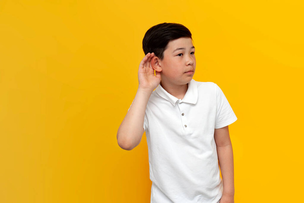 Μικρό ασιατικό αγόρι κρυφακούει σε κίτρινο φόντο, κορεάτικο παιδί κρατά το χέρι κοντά στο αυτί και ακούει σε απομονωμένο φόντο - Φωτογραφία, εικόνα