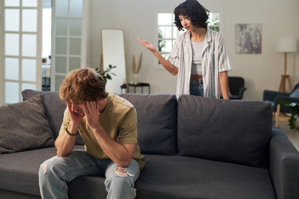 Молодой раздраженный мужчина, сидящий на диване и трогающий его голову, пока его раздраженная жена стоит позади и ругает его во время ссоры - Фото, изображение