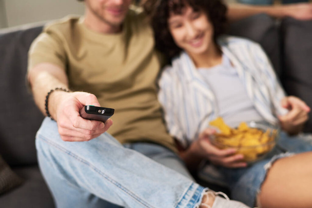 Focus op de hand van de jonge rustgevende man met afstandsbediening tijdens het zitten naast zijn vrouw met chips en het kiezen van tv-kanaal - Foto, afbeelding