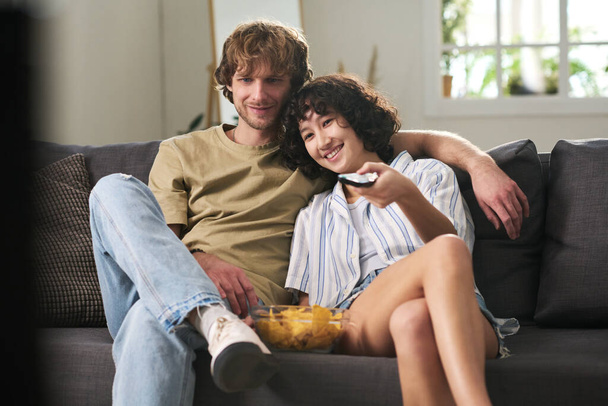 Mladá žena s dálkovým ovládáním a mísa bramborových lupínků výběr zábavný televizní kanál, zatímco sedí na gauči svým manželem - Fotografie, Obrázek