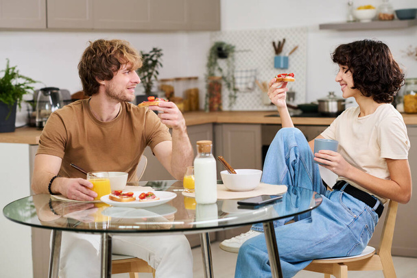 Szczęśliwa młoda para o kanapki warzywne i sok na śniadanie siedząc przy stole w kuchni i omawiając plany na dzień - Zdjęcie, obraz